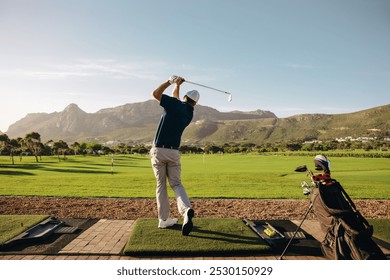 Male golfer taking a swing on a beautiful mountain course at a golf resort, his golf bag standing nearby. Perfect for golf enthusiasts and nature lovers. - Powered by Shutterstock