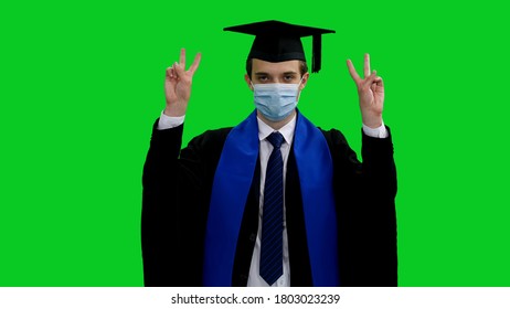 Male Grad Student In Gown And Protective Mask Doing Hand Gesture With Victory Sign Against Green Screen Background, Covid-19 Pandemic, Chroma Key