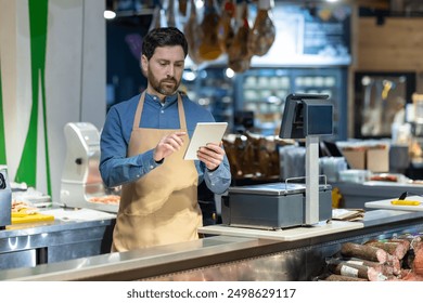 Male grocery store worker using tablet at checkout counter. Worker wearing apron, standing in modern supermarket, checking inventory and prices on device. - Powered by Shutterstock