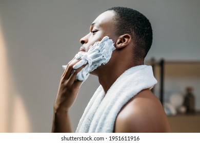 Male grooming. Skincare cosmetology. Morning routine. Profile portrait of confident shirtless masculine African man with white towel on bare shoulders applying shaving foam on dark face skin in light - Powered by Shutterstock