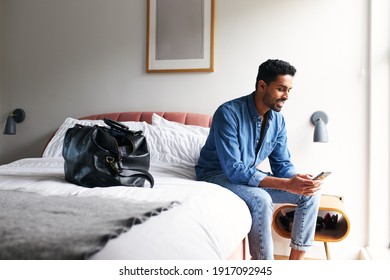 Male guest in boutique hotel sitting on edge of bed checking emails and social media on mobile phone in the morning