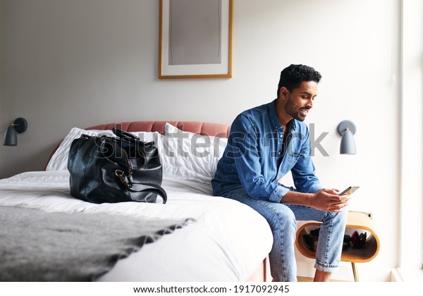 Male guest in\
boutique hotel sitting on edge of bed checking emails and social\
media on mobile phone in the\
morning