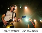 Male guitarist concentrating while playing electric guitar on stage under lights during live concert performance with another band member in background