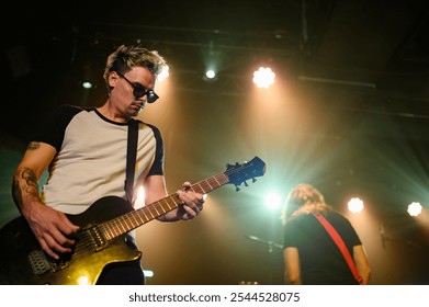 Male guitarist concentrating while playing electric guitar on stage under lights during live concert performance with another band member in background - Powered by Shutterstock