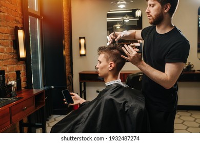 Male Hairdresser Making A Haircut For A Young Man Looking In His Phone In A Barber Shop. He's Standing Behind Client Sitting In A Chair. Side View.