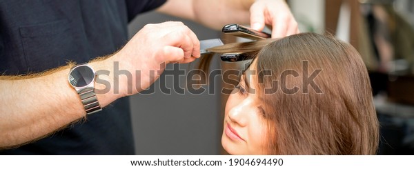 A male hairdresser is straightening the hair\
of the young woman in a beauty\
salon