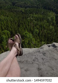 Male Hairy Legs With Sandals Rest On Rocky Summit Above Valley Of Natural Park. Lazy Sunny Summer Day.