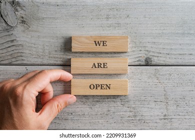 Male Hand Assembling A We Are Open Sign Written On Three Stacked Wooden Blocks Placed Over White Wooden Background.