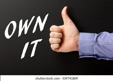 Male hand in a business shirt giving the thumbs up sign to the words Own It written on a blackboard as a reminder to take ownership