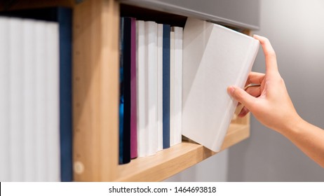 Male hand choosing and picking white book on wooden bookshelf in public library. Education research and self learning in university life concepts
