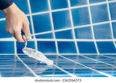 Male hand construction worker using trowel or building spatula spreading cement grout on blue ceramic tiles in the swimming pool