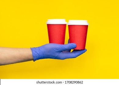 Male Hand Courier Wearing Medical Gloves Holding Paper Cup Coffee For Take Away Isolated On Yellow Background. Contactless Delivery Service In Quarantine Coronavirus Pandemic. Volunteer Make Donation