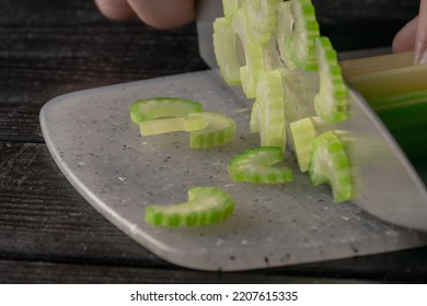 Male Hand Cutting Celery Stalks With A Knife Into Small Slices. Metal Knife Chopping Celery On Gray Plastic Board On Wooden Kitchen Table. Chef Cooking Vegan Salad With Raw Vegetables. Macro Shot.