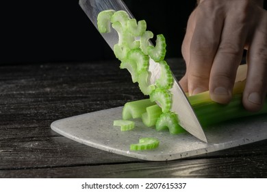 Male Hand Cutting Celery Stalks With A Knife Into Small Slices. Metal Knife Chopping Celery On Gray Plastic Board On Wooden Kitchen Table. Chef Cooking Vegan Salad With Raw Vegetables. Macro Shot.