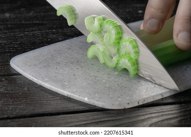 Male Hand Cutting Celery Stalks With A Knife Into Small Slices. Metal Knife Chopping Celery On Gray Plastic Board On Wooden Kitchen Table. Chef Cooking Vegan Salad With Raw Vegetables. Macro Shot.