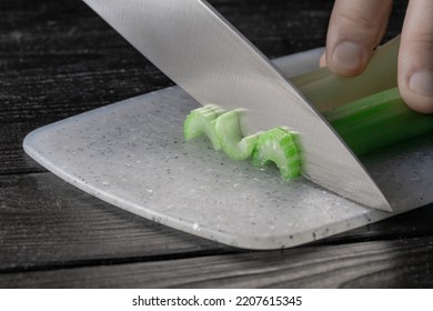 Male Hand Cutting Celery Stalks With A Knife Into Small Slices. Metal Knife Chopping Celery On Gray Plastic Board On Wooden Kitchen Table. Chef Cooking Vegan Salad With Raw Vegetables. Macro Shot.