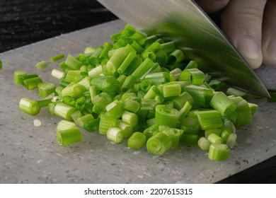 Male Hand Cutting Green Onion Stalks With Knife Into Small Slices. Metal Knife Chopping Shallots Or Chive On Gray Plastic Board On Wooden Restaurant Kitchen Table. Chef Cooking Vegan Salad. Macro Shot