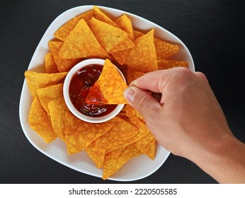 male hand dipping a mexican corn chip or tortilla chip in a spicy salsa dip, top view of a bowl of tortillas with salsa on black slate background - Powered by Shutterstock