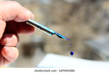 In The Male Hand Is A Fountain Pen. A Drop Of Blue Ink Drops From A Metal Pen. Blurred Background. Concept - Education