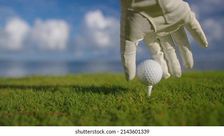 Male Hand In A Golf Glove Puts The Ball On The Field On The Background Of The Blue Ocean. Close Up