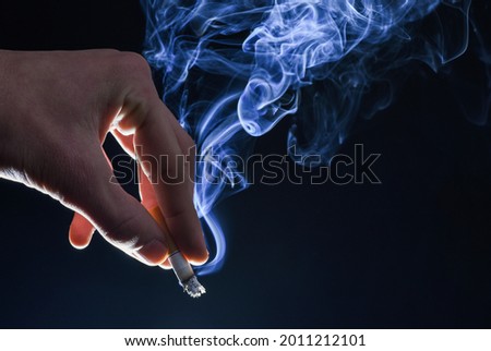 Male hand hold cigarette butt with smoke swirls dark background, nicotine addiction