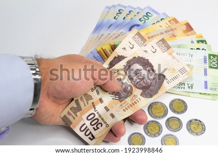 Male hand holding 500 Mexican peso bills on top of a pile of other bills and coins of various denominations.