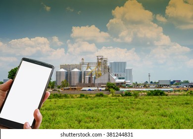 Male Hand Holding Blank Smart Phone And Building Exterior, Factory Of Agriculture.