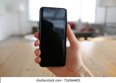 A Male Hand Holding A Brand New, Very Modern, State Of The Art Mobile Phone. The Touch Screen Is Black With Reflections On It. An Image On An Interior Design Background With A Visible Wood Table.