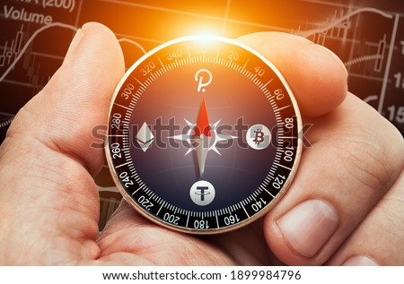 Male hand holding compass with bitcoin, ethereum, polkadot and tether logo in front of stock market chart data. Compass needle showing  polcadot coin symbol.