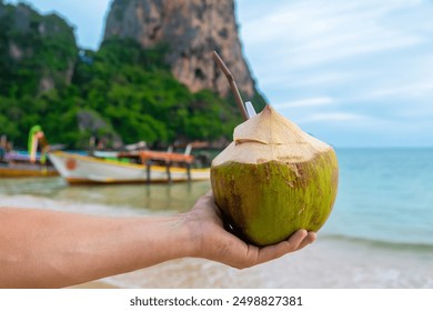 Male hand holding fresh coconut drink on tropical sandy Railay beach with cliffs and traditional Thai boats at Krabi province, Thailand. Man holds refreshment summer drink with a straw - Powered by Shutterstock