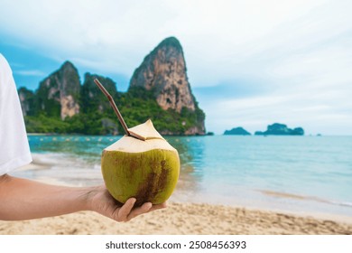 Male hand holding fresh coconut drink on tropical sandy Railay beach with cliffs at Krabi province, Thailand. Man holds refreshment summer drink with a straw on the sea coast. Close up - Powered by Shutterstock