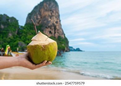 Male hand holding fresh coconut drink on tropical sandy Railay beach with cliffs and traditional Thai boats at Krabi province, Thailand. Man holds refreshment summer drink with a straw - Powered by Shutterstock