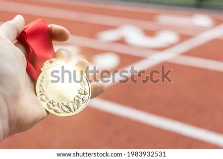 Male hand holding a gold medal in front of a running track.