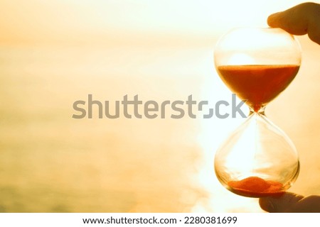 male hand holding Hourglass at sunset sea background with empty copy space. sun set sky. time is money. 