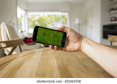 A Male Hand Holding A Mobile Phone Which Displays A Soccer Match On The Touch Screen. An Image On An Interior Background.