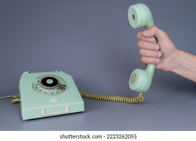 Male hand holding an old blue plastic telephone receiver near rotary telephone on gray background. Close up remote handset from retro home phone apparatus in the hands of man.