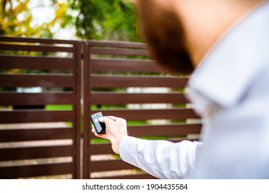 A male hand holding a remote control to open an entrance gate. An automatic opening, closing of a driveway gate. A modern house.