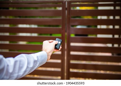 A Male Hand Holding A Remote Control And Pushing A Button To Open An Entrance Gate. An Automatic Opening, Closing Of A Driveway Gate. A Modern House.