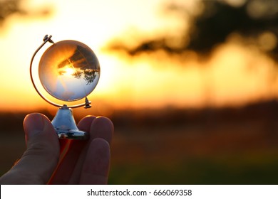 Male Hand Holding Small Crystal Globe In Front Of Sunset. Travel And Global Issues Concept