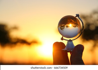 Male Hand Holding Small Crystal Globe In Front Of Sunset. Travel And Global Issues Concept