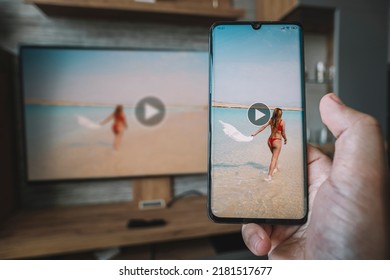 Male Hand Holding A Smartphone Against View Of Living Room With A Large Screen TV. Use Wi Fi To Transfer Images From Phone On The Monitor Screen. Viewing Video From Your Smartphone On Your TV.