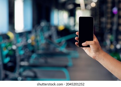 Male Hand Holding Smartphone With Blank Black Screen Over Gym Interior Background, Unrecognizable Athlete Showing Template With Free Copy Space For Fitness App Or Website Advertisement, Mockup