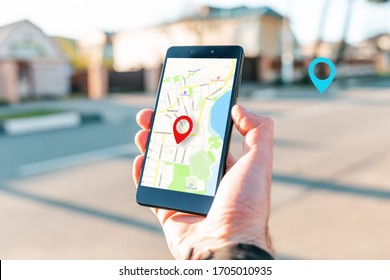 Male hand holding holding smartphone with online-card, which is a red icon of the location. In the background, there is a blurred street and a blue location icon. Concept of online navigation and GPS