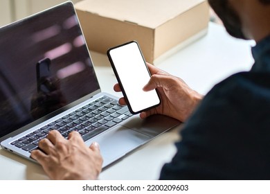 Male hand holding smartphone with white mock up screen using laptop at work desk. Business man showing blank mobile display mockup template of cell phone with empty space, over shoulder view.
