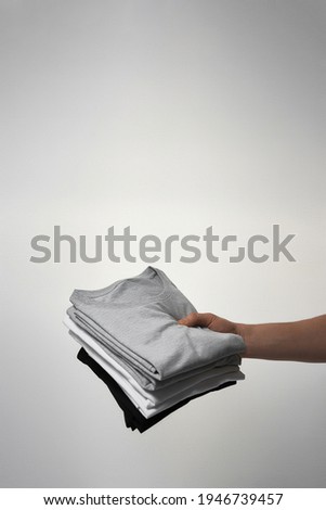 Male hand holding stack of basic mens T-shirts against gray background. Casual clothes for people, fashion concept