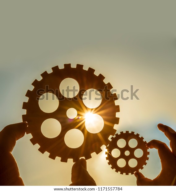 Male hand\
holding two iron cog gear wheel on sunny sky background. sunshine\
and yellow and blue sky . sun\
rays.