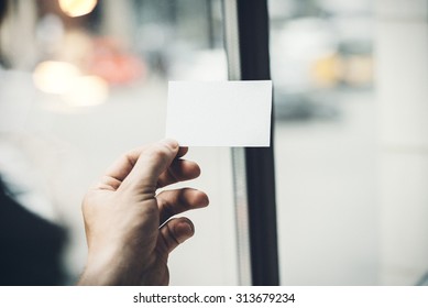 Male Hand Holding White Business Card On The Background Of City 