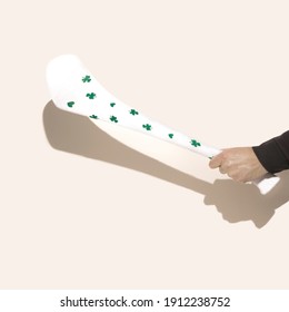 A Male Hand Holding White Hurling Stick, Hurley With  With Shamrock Decoration. Beige Background.