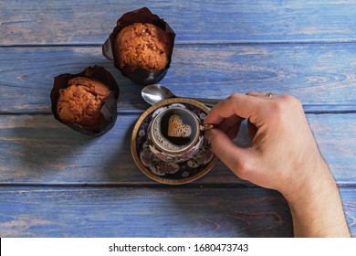 Male hand holds a cup of black coffee with foam in the shape of a heart and vanilla muffins on a background of blue wooden table