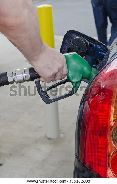 Male hand holds a pump nozzle filling up car with petrol\
at a petrol station. Car tail light and petrol filling door on\
image. Car is dark blue and petrol nozzle is green. Vertical\
portrait image. 